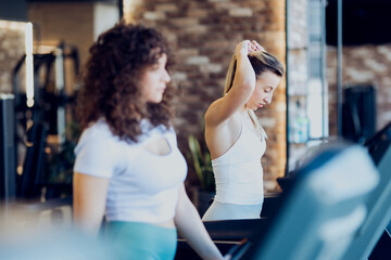 Two young women in athletic wear preparing to start their workout on treadmills in a modern gym. They are adjusting the settings and getting ready to run
