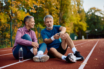 Mature sports couple using fitness tracker while relaxing on running track outdoors.