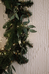 Evergreen Christmas garland with pearl and gold ornaments decorating white fireplace against beige wall. Warm fairy lights create festive atmosphere. Concept for holiday decor, winter, celebration.