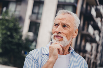 Elderly man in casual striped shirt stands on a sunny urban street thoughtfully looking into the...