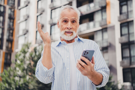 Elderly man in striped shirt uses smartphone outdoors in a modern city setting waving hand as he chats during a casual day by apartment buildings