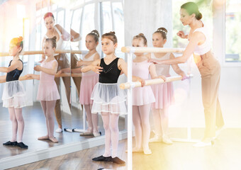 Naklejka premium Adult female teacher corrects group of girls dancers standing in first position at ballet barre