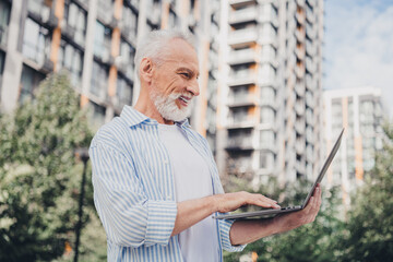 Senior man outdoors in the city using a laptop smiling relaxed urban day beside modern residential buildings