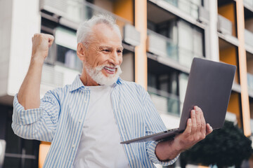 Obraz premium Elderly man outdoors in a modern city holds a laptop and smiles at the screen celebrating success and connection