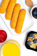 Healthy continental breakfast with corn sticks, granola with blueberries, orange juice, and red fruit and cherry jams on a white background.