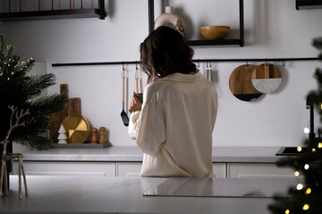 Woman in cream satin pajamas in the kitchen holds a cup of coffee in the morning Christmas. Preparation for winter holidays and New Year Eve. Happy young woman having fun and dreaming.