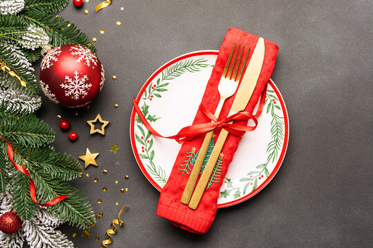Festive Christmas table setting with white ceramic plate and golden cutlery set on dark stone background decorated with fir tree branches and red balls.