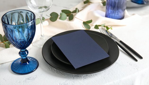 Stylish table setup with black dinnerware, blue goblet, and empty navy card on a white textured background, decorated with eucalyptus leaves, for event planning or dining