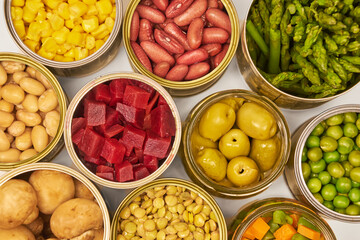 Open cans of beans, mushrooms, carrots, corn, peas, green beans, lentils, asparagus, and olives on a white background with copy space.