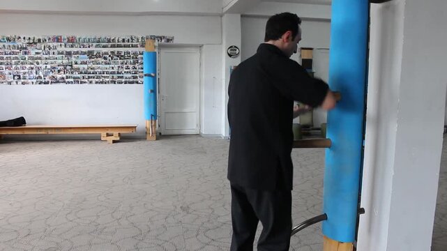 A professional martial artist wearing a black uniform is practicing Wing Chun techniques on a blue wooden dummy inside a traditional Chinese martial arts training studio.


