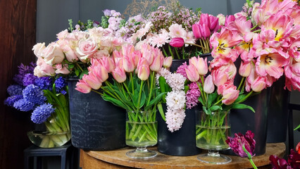 Colorful assortment of fresh flowers displayed in black buckets inside flower shop. Various blooms including tulips, roses, mixed spring flowers arranged on and around a wooden table on floral market 