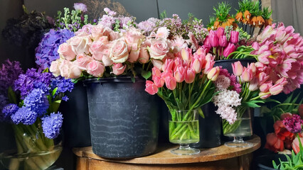 Colorful assortment of fresh flowers displayed in black buckets inside flower shop. Various blooms including tulips, roses, mixed spring flowers arranged on and around a wooden table on floral market 