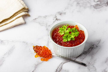Bowl of red caviar with parsley and spoon on marble background