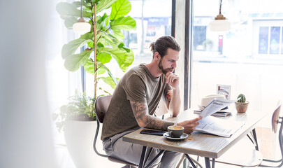 Tattooed man deeply focused on reading newspaper while sitting in a bright modern café with coffee and a smartphone.