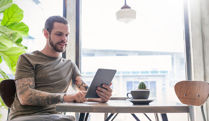 Young tattooed man sitting at a modern cafe, using a tablet near a window with coffee and a small cactus nearby. Relax slow life with technology