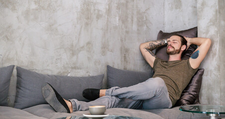 Young man with tattoos resting on a stylish gray couch in an urban apartment, enjoying a peaceful and calm moment.