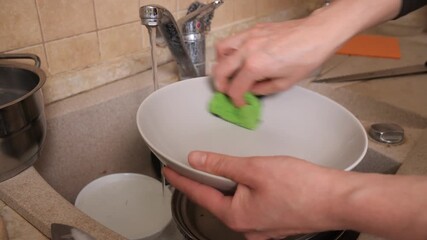 Person washing a plate with a green sponge under running kitchen faucet, scrubbing away food and soap suds in sink as part of daily household cleaning routine and hygiene maintenance