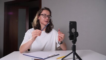 Woman explaining art techniques during a remote, live streamed education session, sitting at a desk and holding pencils while recording on a smartphone