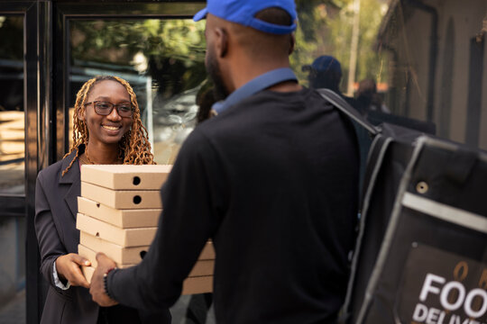 Delivery man brings a stack of pizza boxes to a woman standing at front door. Quick and friendly interaction between african american people for a good delivery experience with huge order.