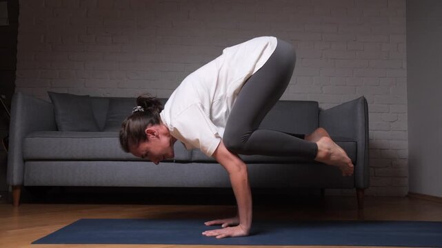 Woman balancing in bakasana yoga pose on a blue mat, performing an arm balance exercise for strength, focus, and mindfulness during a home workout session