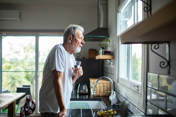 Senior man sipping water, thoughtful at home kitchen window