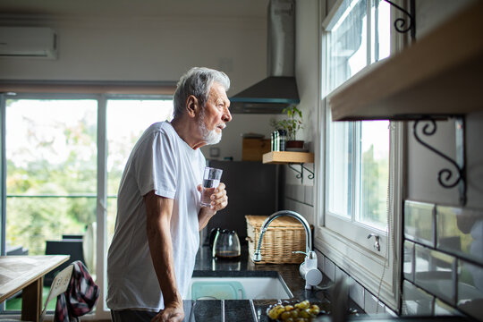 Senior man sipping water, thoughtful at home kitchen window