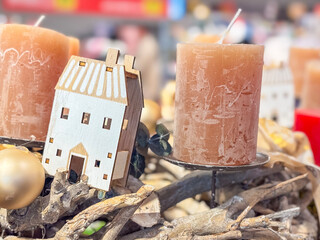 close-up of wooden miniature house decoration between rustic orange pillar candles and gold bauble on branches concept of cosy home decor, winter greetings, holiday gifts