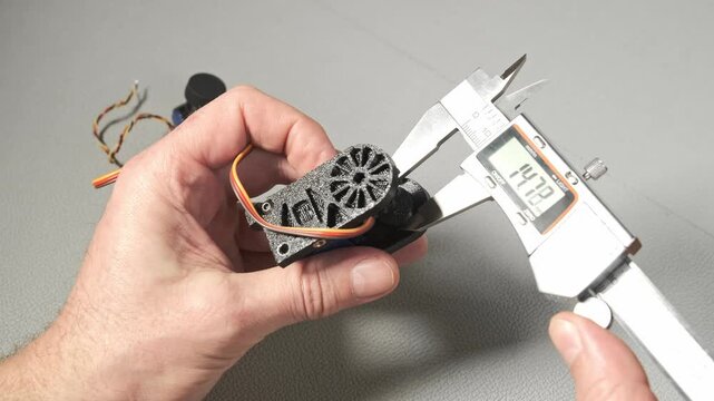 An engineer works with a digital caliper to measure a small robotic servo component with gears and wiring. Taking size, wide, length The precise measurement is shown on the LCD screen of the tool.