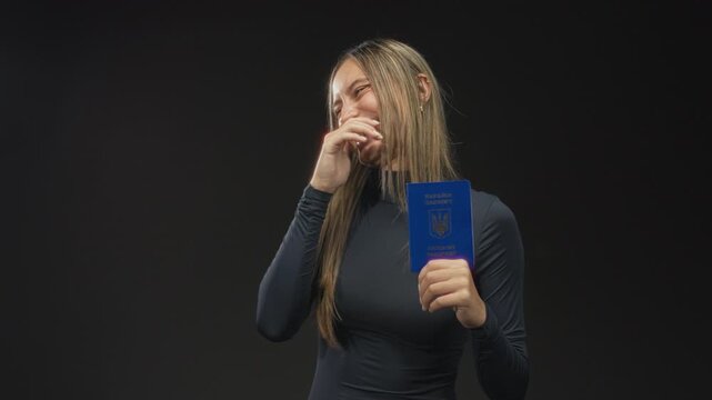 Woman holding ukraine passport with hand covering mouth while laughing and showing passport front in studio black backdrop; joy travel.