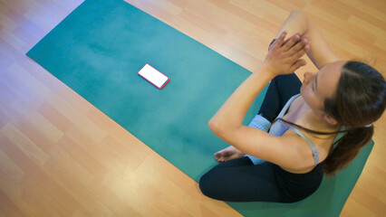 A woman sits on a green yoga mat on a wooden studio floor, performing a seated stretch with hands...