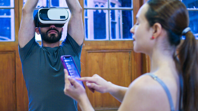 A man wears a virtual reality headset with arms raised, immersed in a digital experience, as another woman nearby interacts with a smartphone, suggesting tech collaboration.
