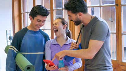 Three friends share a joyful moment during a workout in a bright studio; one holds a phone while another adjusts a trainer’s stance, creating a lively, energetic fitness scene.
