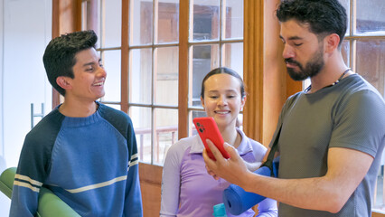A group of three young adults indoors by large windows, smiling and focused on a red smartphone. Casual outfits and a relaxed vibe capture a moment of connection