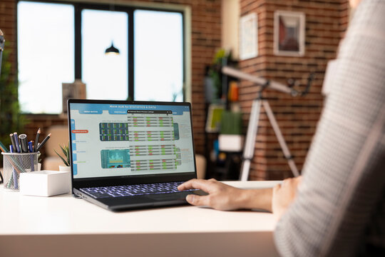 Remote working woman reviewing financial reports on laptop at home office desk. Female business analyst sitting and monitoring company spending in stylish brick wall apartment.