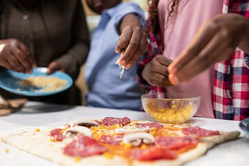 Close up of black young kids adding grated mozzarella on pizza dough, sharing pizza day at home and learning to cook with their parents for bonding time. Togetherness and laughter.