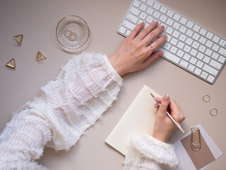 A simple, feminine workspace with a beige background, featuring a woman's hands typing on a wireless keyboard while taking notes, a woman's computer desk, and a flat lay top view of her hands at work.