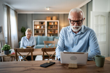 Senior man using digital tablet at home while wife reads
