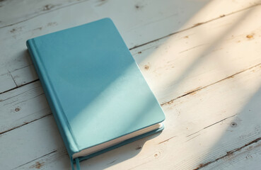 Light blue notebook with plain cover lies on distressed white wood table. Sunlight casts soft shadows across surface. Blank journal ready for new ideas, planning, school study, work notes, daily