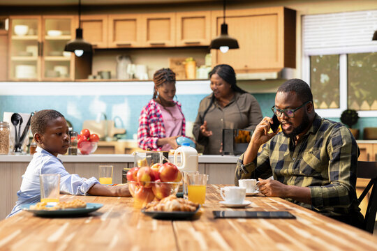 African american father interrupting breakfast meal to answer a phone call, neglecting his family and being busy at home. Working dad ignoring son and daughter at the table.