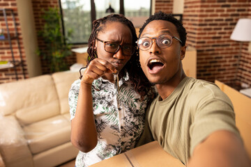 Young african american couple share unpacking humor in new apartment living room. Black woman proudly holds house keys as boyfriend balances box, taking fun selfie together during moving day.