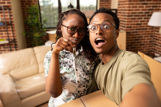 Young african american couple share unpacking humor in new apartment living room. Black woman proudly holds house keys as boyfriend balances box, taking fun selfie together during moving day.