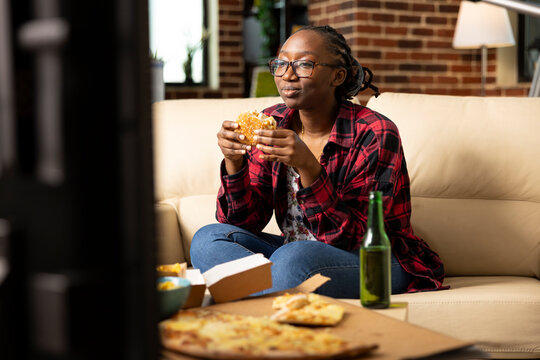 Smiling black woman relaxing on couch, grasping burger and and watching tv show with concentration. Young lady spends time at home enjoying cozy entertainment, takeaway meal and peaceful downtime.