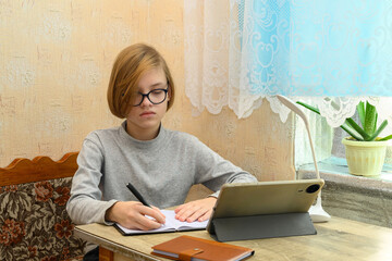 boy in glasses studying at a table, writing in a notebook with a tablet and desk lamp nearby