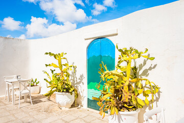 Bright courtyard with white walls, cacti, and a turquoise arched door under blue skies in Ostuni town, Apulia, Italy