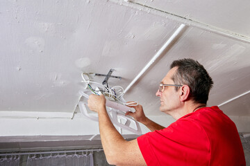 Mounting electrician lifts LED chandelier to ceiling in rural house, wires positioned for connection, fixture aligned with bracket.