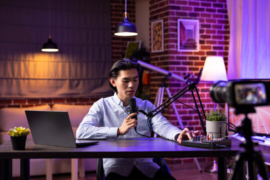 Focused asian man sets up audio equipment in modern home studio, seated at desk checking sound on mixer. Male podcaster uses microphone to speak to online audience during digital talk show broadcast.