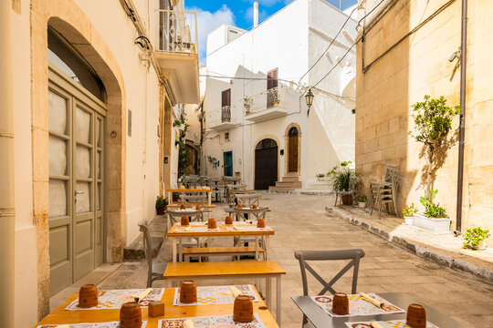 Sunlit narrow street with outdoor cafe tables and people enjoying lunch by old stone walls in Ostuni town, Apulia, Italy