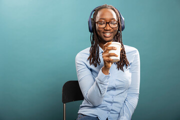 Black female individual with glasses, seated on chair, listening to music and gazing at her coffee cup. Smiling african american woman wearing wireless headphones and enjoying hot beverage.