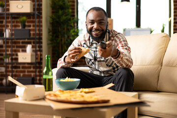 Happy black man holds pizza slice and remote control, increasing TV volume while enjoying good movie. Relaxed weekend moment in modern brick wall apartment, full of comfort and joy.