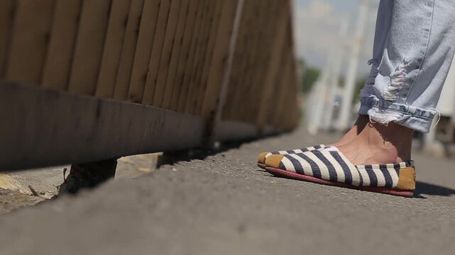 Close up of feet in striped slip-on shoes and distressed jeans resting on a wooden fence rail against a blurry background on a sunny day.


